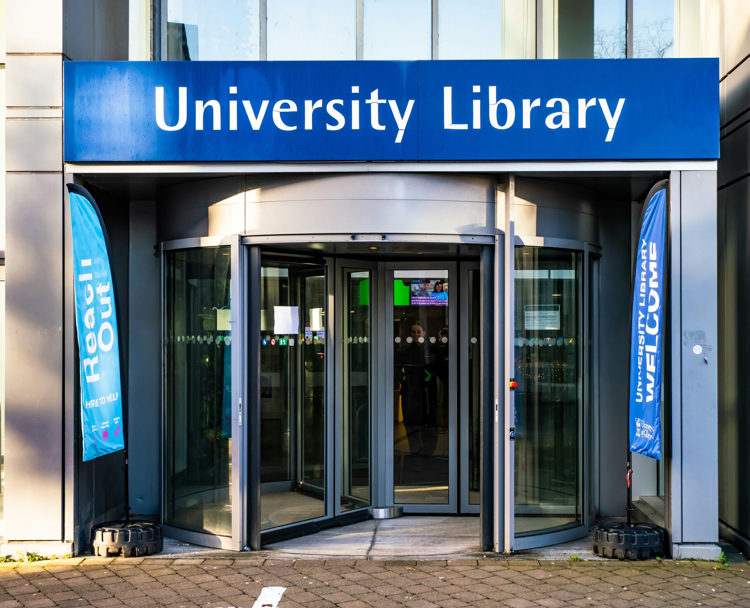 A photograph of the doors to a university library on a sunny day. Above the revolving doors the words University Library are written in white against a blue background.